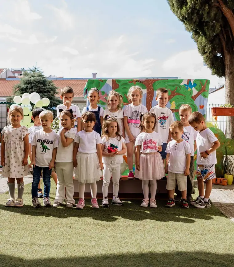 Grupna fotografija oko petnaestoro dece u belom, poređanih u dva reda ispred ručno oslikane kulise sa drvetom i pticama