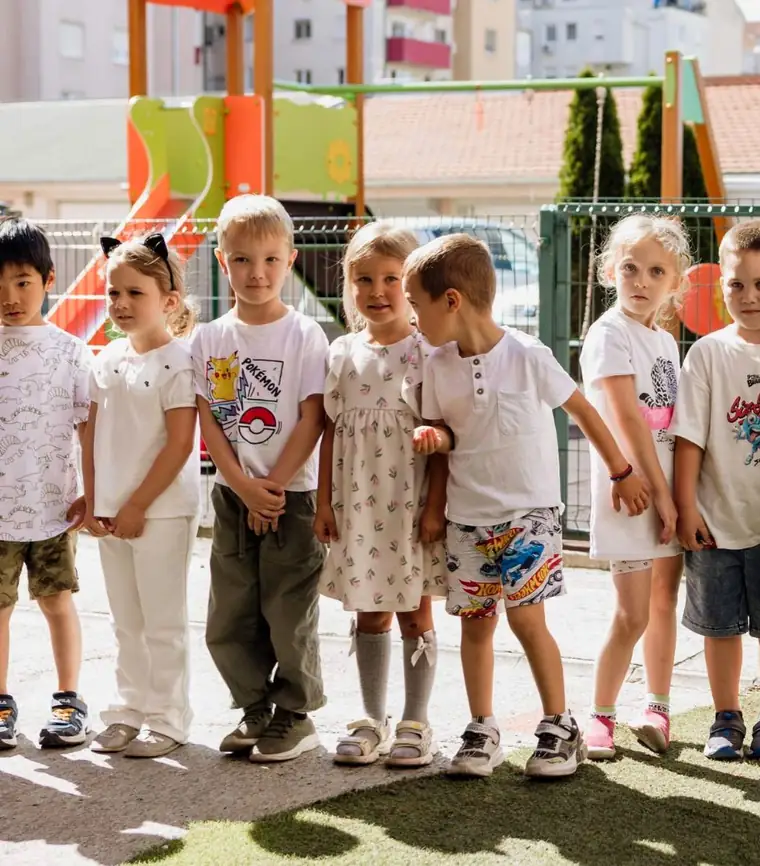 Sedmoro dece stoji u redu na betonskoj stazi ispred dvorišnog igrališta, u sunčano letnje popodne